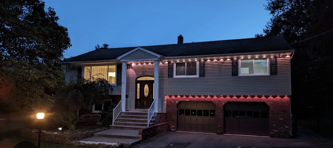 A split level home at dusk lit by a boxed consumer light kit, showing repeating bright wedges and dark gaps along the eave and above the garage