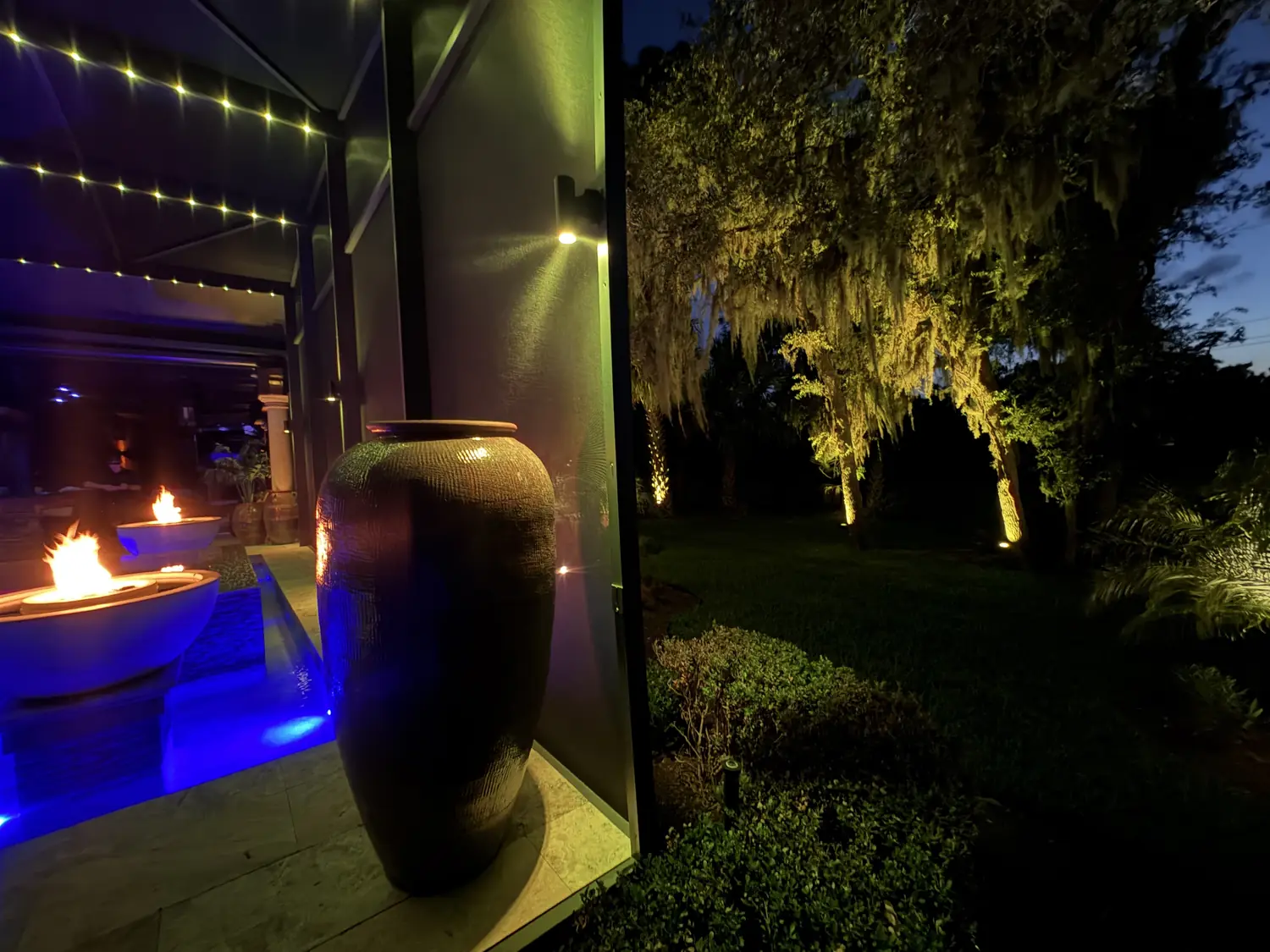 Pool enclosure sconces and landscape lighting in warm white at dusk, fire bowls burning and live oaks lit beyond the screen