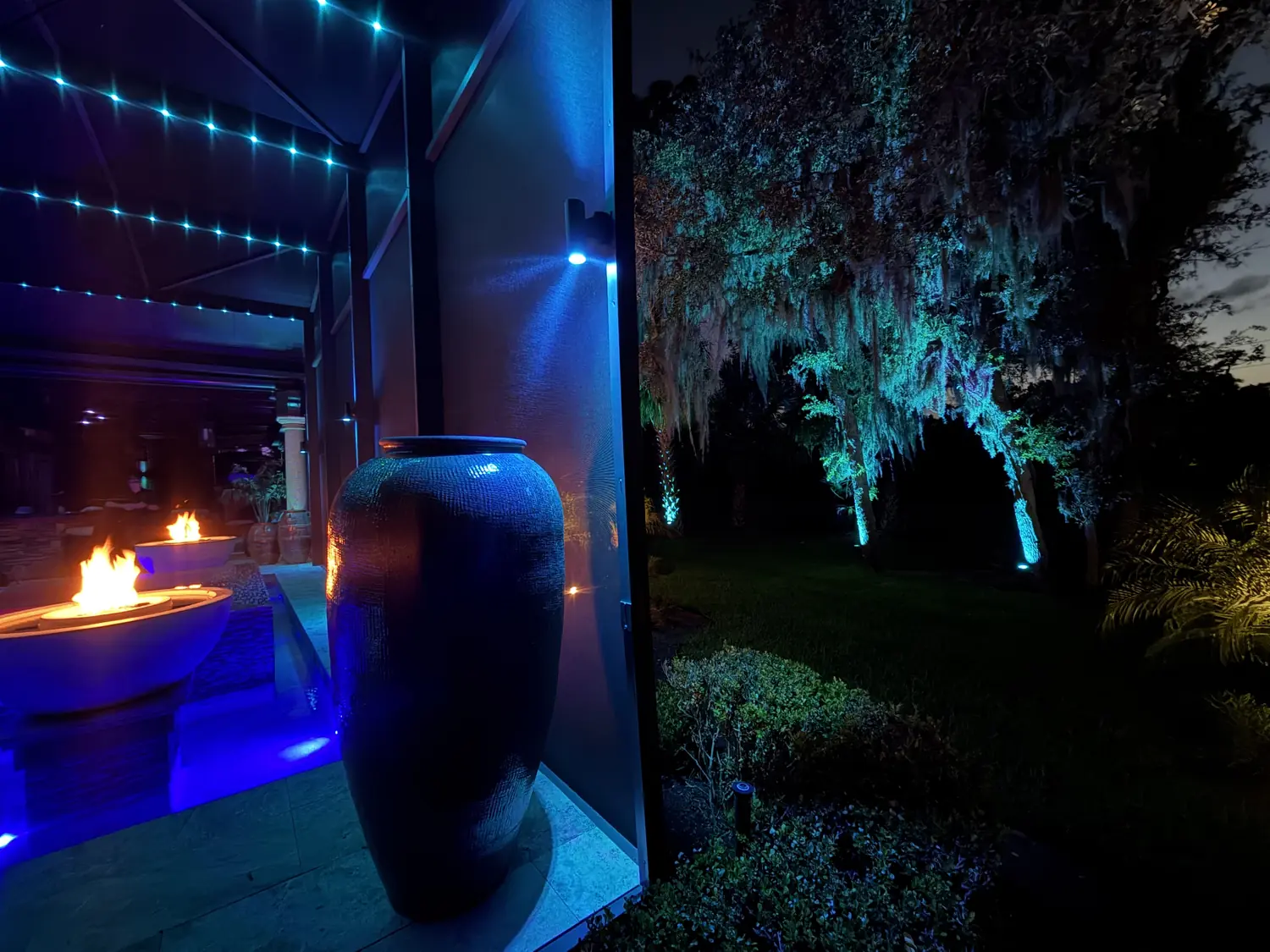 Pool enclosure sconces and tree uplights in ocean blue, fire bowls burning against the cool wash